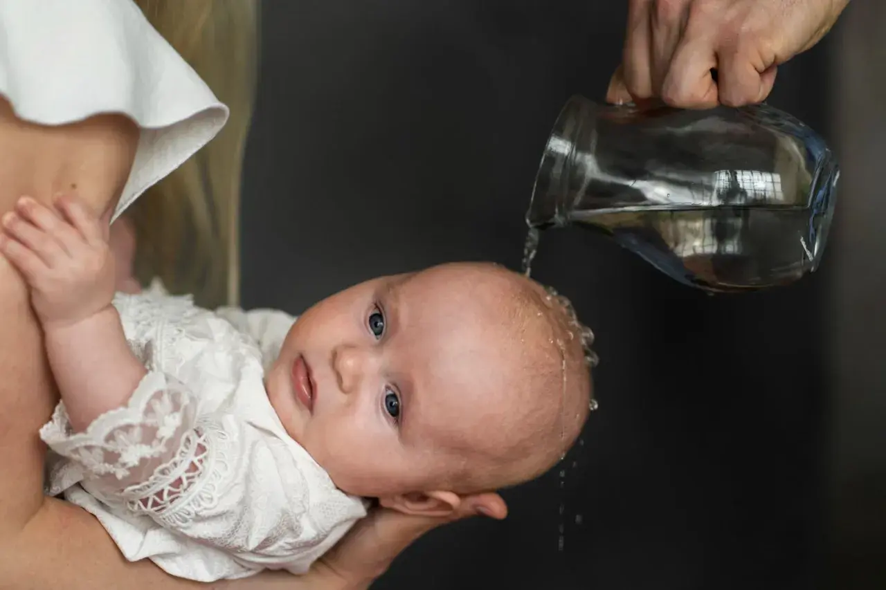 Chrzest dziecka. Woda spływa po głowie malucha w białej szatce. Niech to będzie początek mądrych życzeń na chrzest.