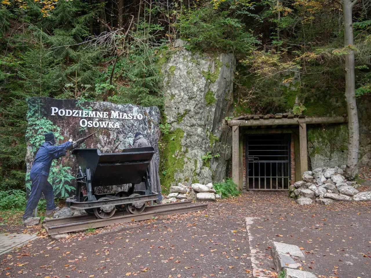 Wejście do Podziemnego Miasta Osówka, z wagonikiem i tablicą informacyjną. Widać skalne ściany i wejście do tunelu.