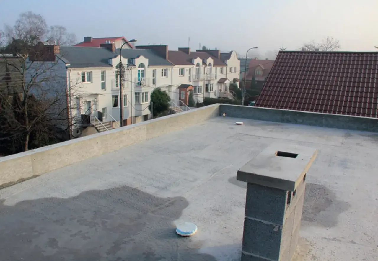 Nowy, betonowy dach płaski z kominem i widokiem na szereg domów z dachówką.