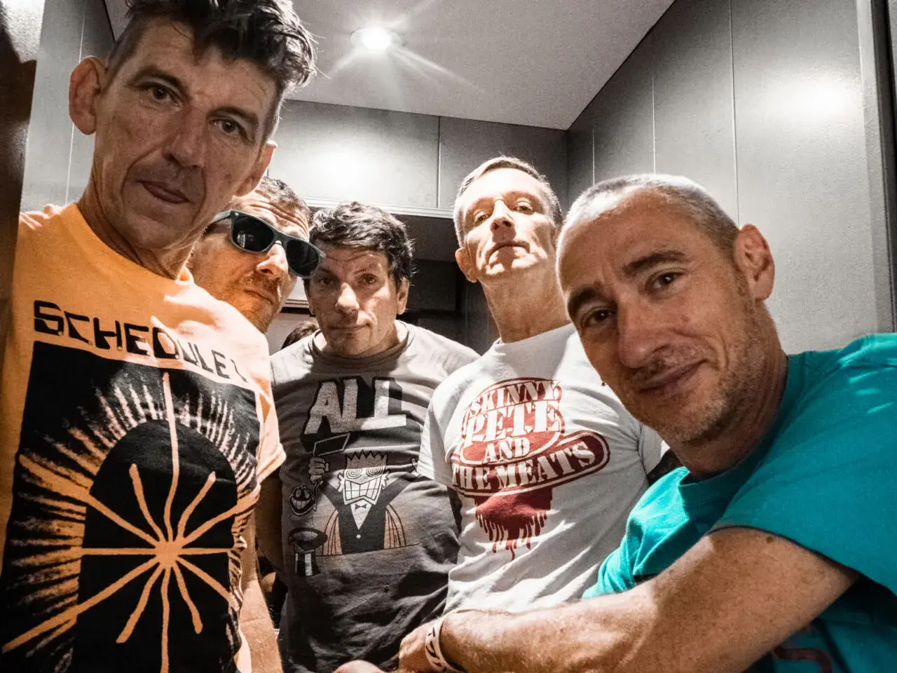 Cinq hommes aux têtes brûlantes, certains souriants, d'autres plus intenses, posent pour un selfie. Leurs t-shirts affichent des graphismes variés.