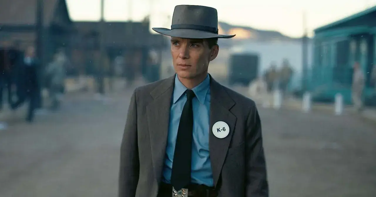 Julius Robert Oppenheimer w kapeluszu i garniturze, z odznaką K-6, w scenerii przypominającej miasteczko.