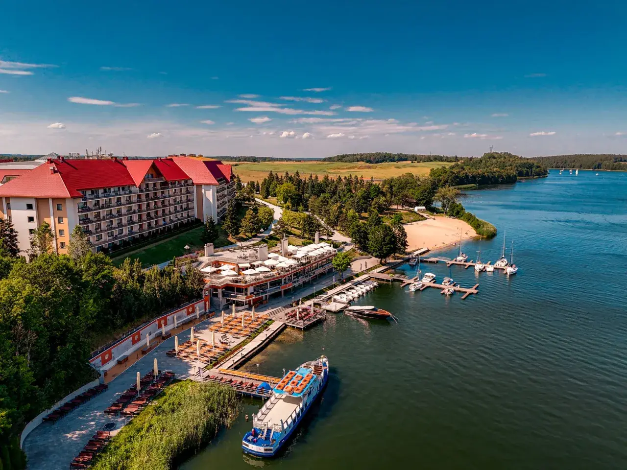 Hotel na wyspie Mikołajki z czerwonym dachem, otoczony zielenią, z mariną pełną łodzi i statkiem wycieczkowym na jeziorze.