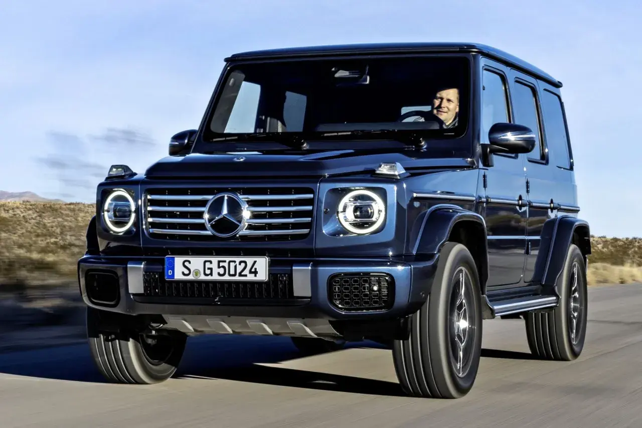 Ciemnoniebieski Mercedes G Klasa jedzie po pustynnej drodze. Kierowca uśmiecha się. Sprawdź mercedes g klasa cena!