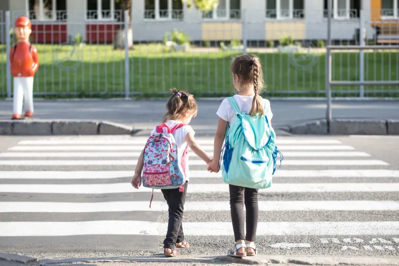 Dwie dziewczynki z plecakami idą za rękę przez pasy. Od ilu lat dziecko może samo chodzić po mieście?