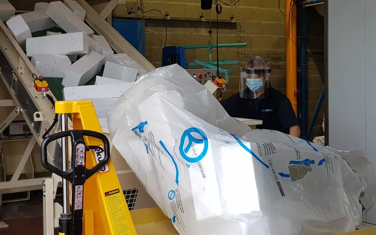Pracownik w masce i przyłbicy przygotowuje styropian do recyklingu. Duży worek z logo Knauf Circular symbolizuje proces recyklingu styropianu.