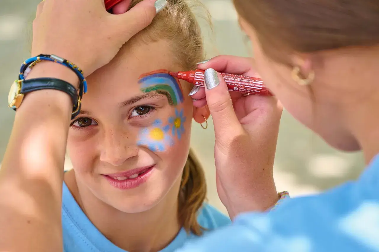 Une petite fille souriante se fait maquiller sur la joue un arc-en-ciel et des fleurs, au stand maquillage d'une kermesse.