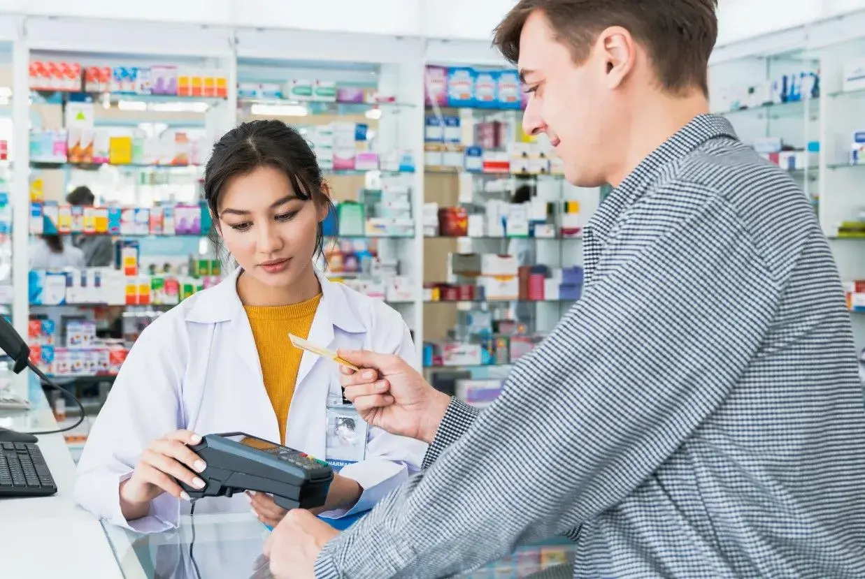 Klient płaci kartą w aptece, gdzie farmaceutka obsługuje terminal. W tle widać półki z lekami, a na nich produkt z napisem ursopol.