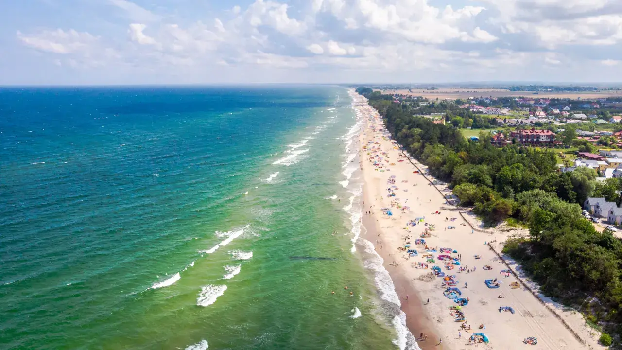 Słoneczny dzień na plaży w Sarbinowie nad morzem. Tłumy turystów odpoczywają na piasku, kąpią się w morzu i spacerują wzdłuż brzegu.