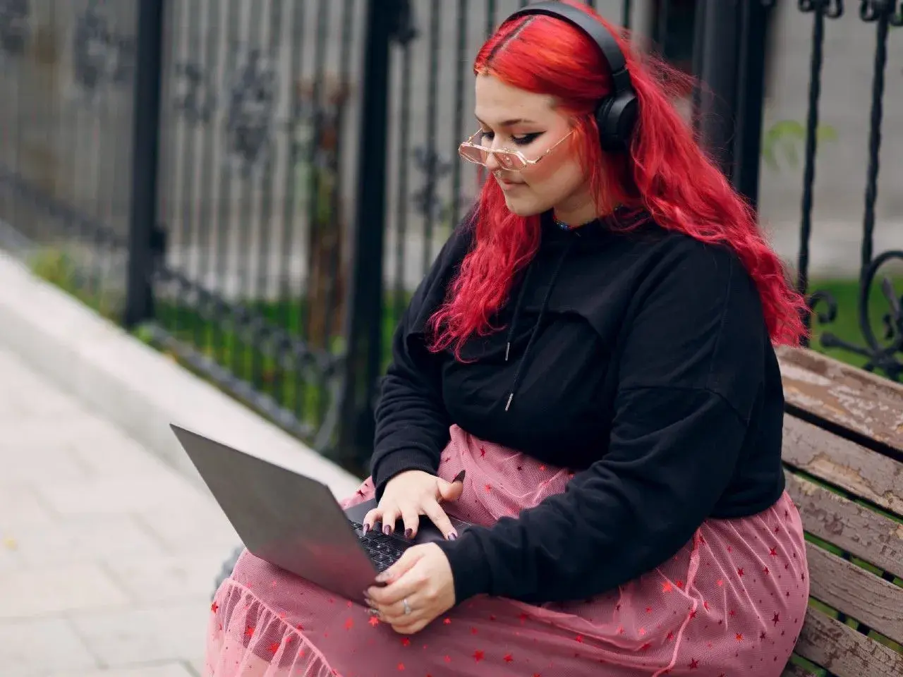 Młoda babista z ognistymi włosami i słuchawkami na uszach pracuje na laptopie, siedząc na ławce w parku.
