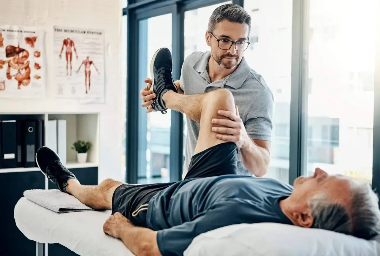 Ein Physiotherapeut dehnt das Bein eines Patienten. Der Patient liegt auf einer Liege, während der Therapeut sein Knie beugt. Wann Physiotherapie? Hier wird geholfen.