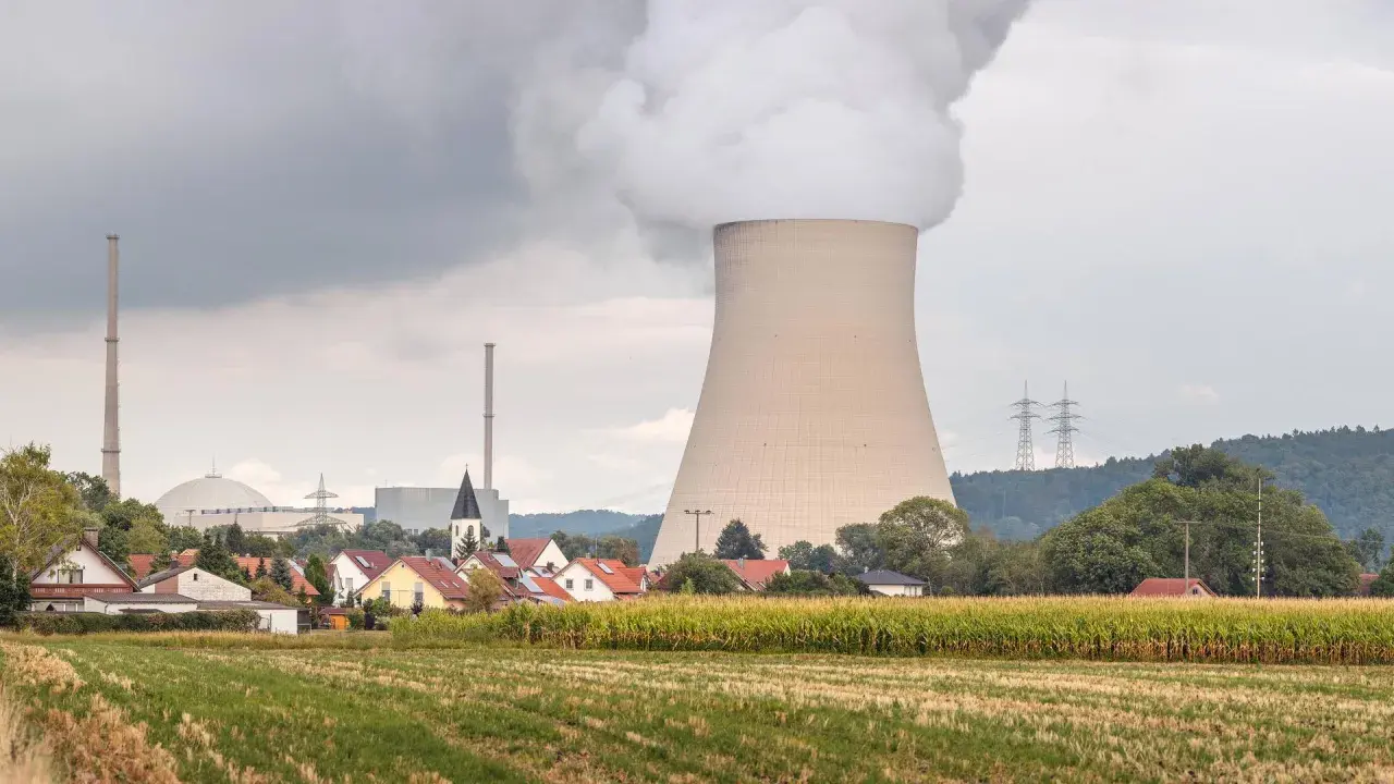 Dampf steigt aus einem Kühlturm eines Atomkraftwerks auf, daneben ein Dorf und ein Maisfeld.