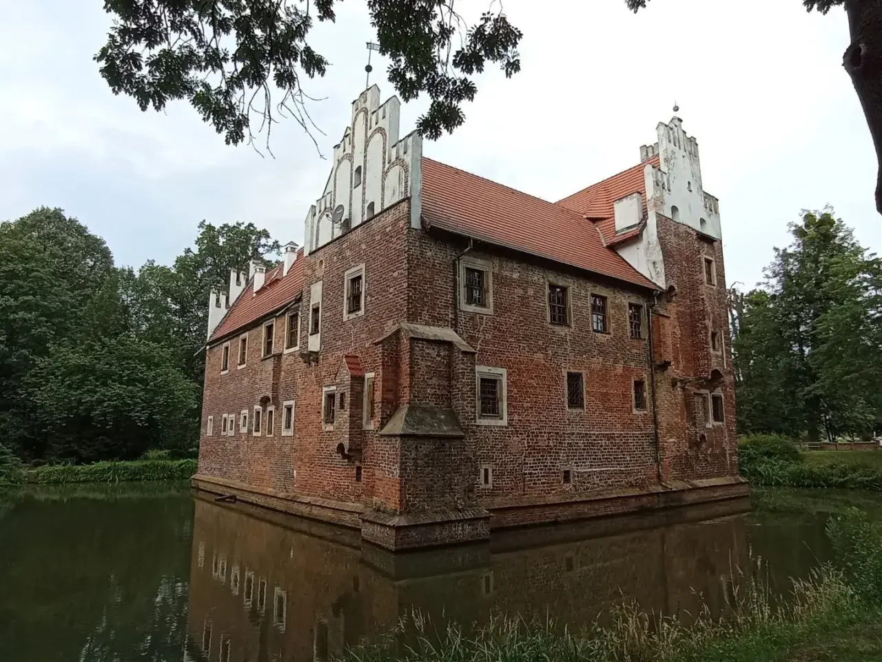 Zamek na wodzie w Wojnowicach, otoczony fosą, prezentuje ceglaną architekturę z białymi zdobieniami.