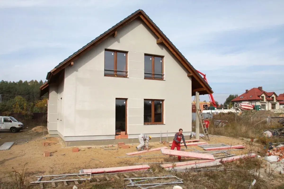 Budowa domu w toku. Pracownicy układają płyty, a w tle widać betonowóz. Czas budowy zależy od wielu czynników.