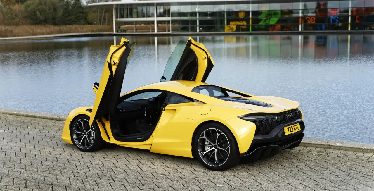 Żółty McLaren Artura z otwartymi drzwiami, stojący nad jeziorem przy nowoczesnym budynku.