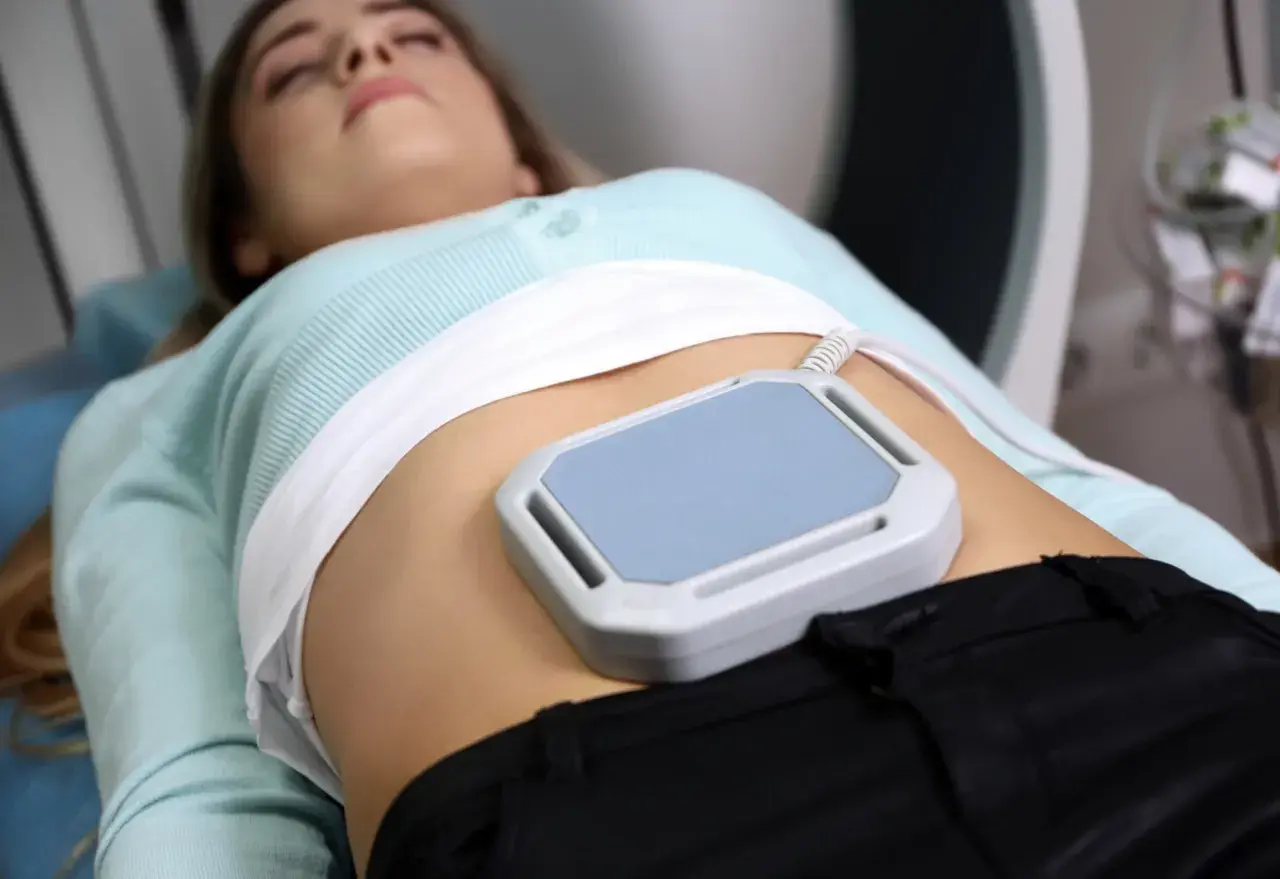 Kobieta leży podczas zabiegu, na brzuchu ma nałożone urządzenie do terapii polem magnetycznym, co jest częścią jej rehabilitacji.