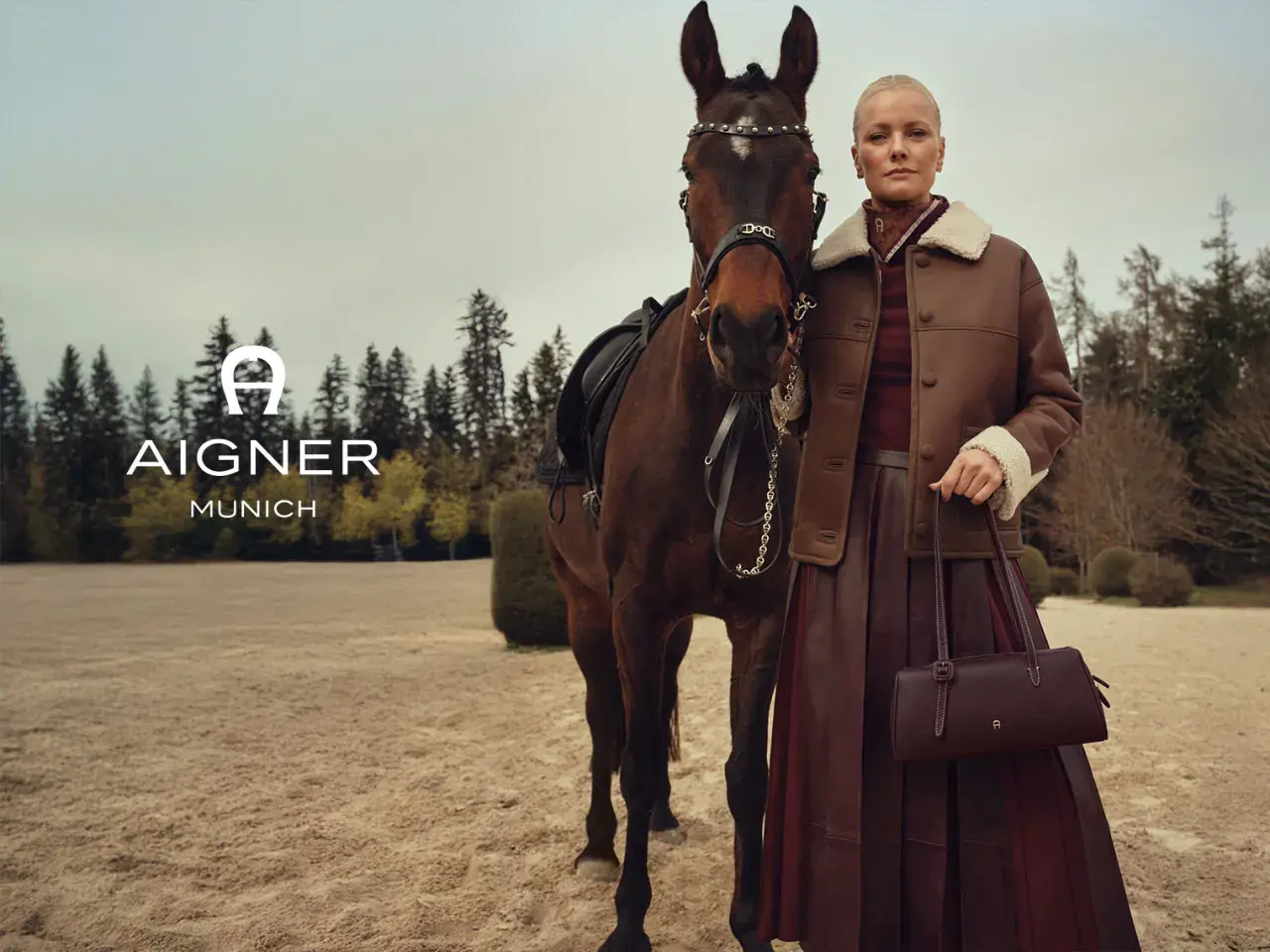 Elegancka kobieta w brązowej kurtce i spódnicy obok konia. Logo AIGNER MUNICH. Aigner co to za marka? Luksusowa moda.