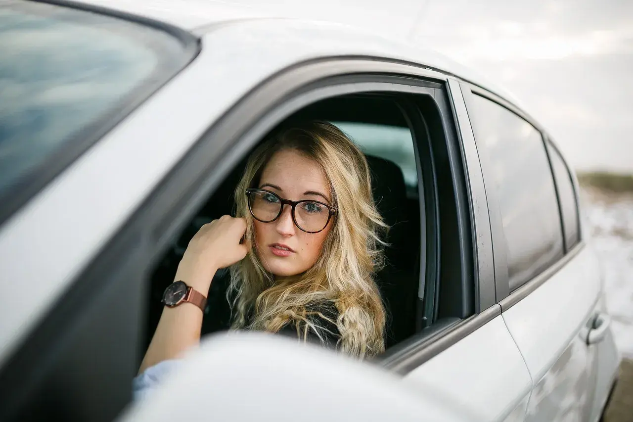 Blondynka w okularach siedzi za kierownicą białego samochodu. Pokazuje, jak nosić okulary, by wyglądać stylowo i pewnie.