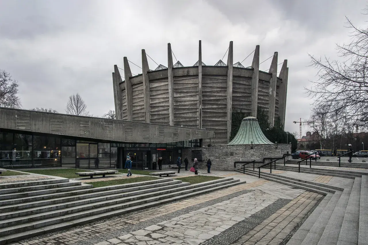 Monumentalny budynek w stylu brutalizm z betonowymi żebrami i szklaną fasadą. Schody prowadzą do wejścia, a wokół zielone trawniki.