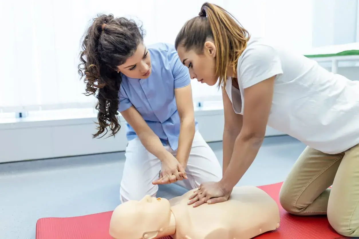 Zwei Frauen üben kardiopulmonale Reanimation an einer Übungspuppe. Eine Frau gibt Anweisungen, während die andere die Herzdruckmassage durchführt.