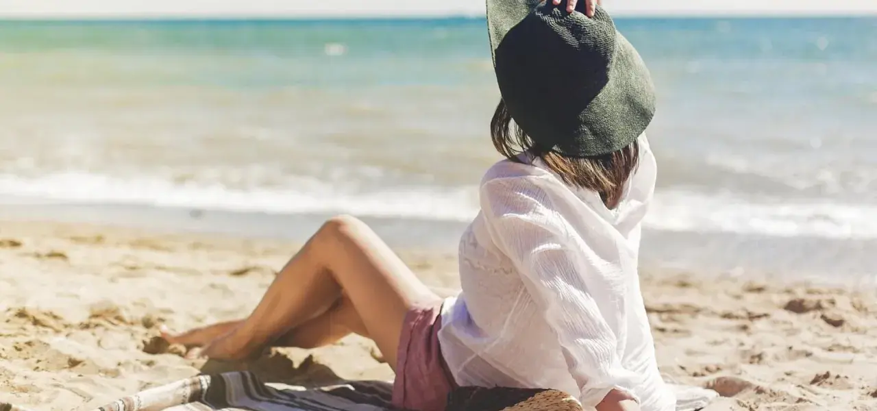 Kobieta w kapeluszu siedzi na plaży, w tle widać spokojne jezioro. Idealne miejsce na ustronny domek nad jeziorem.