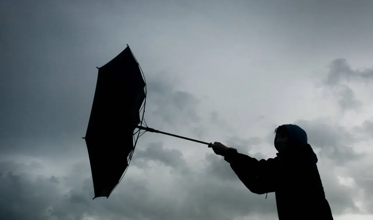 Silhouette einer Person mit einem aufgewehten Regenschirm vor dramatischem Himmel. Der **Orkan** kämpft gegen den Schirm.