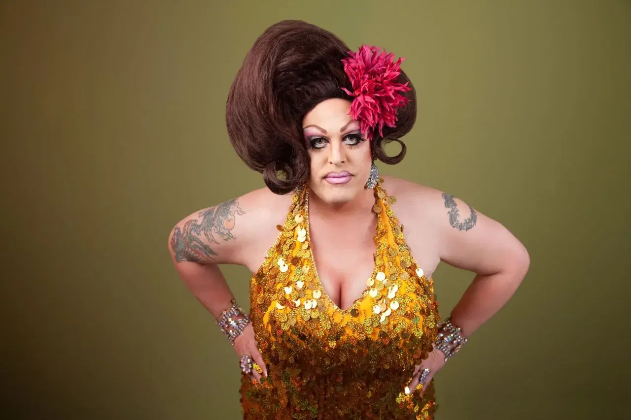 Osoba w złotej, cekinowej sukience i peruce, z kwiatem we włosach. To przykład tego, co to jest drag queen – artystyczna forma ekspresji.