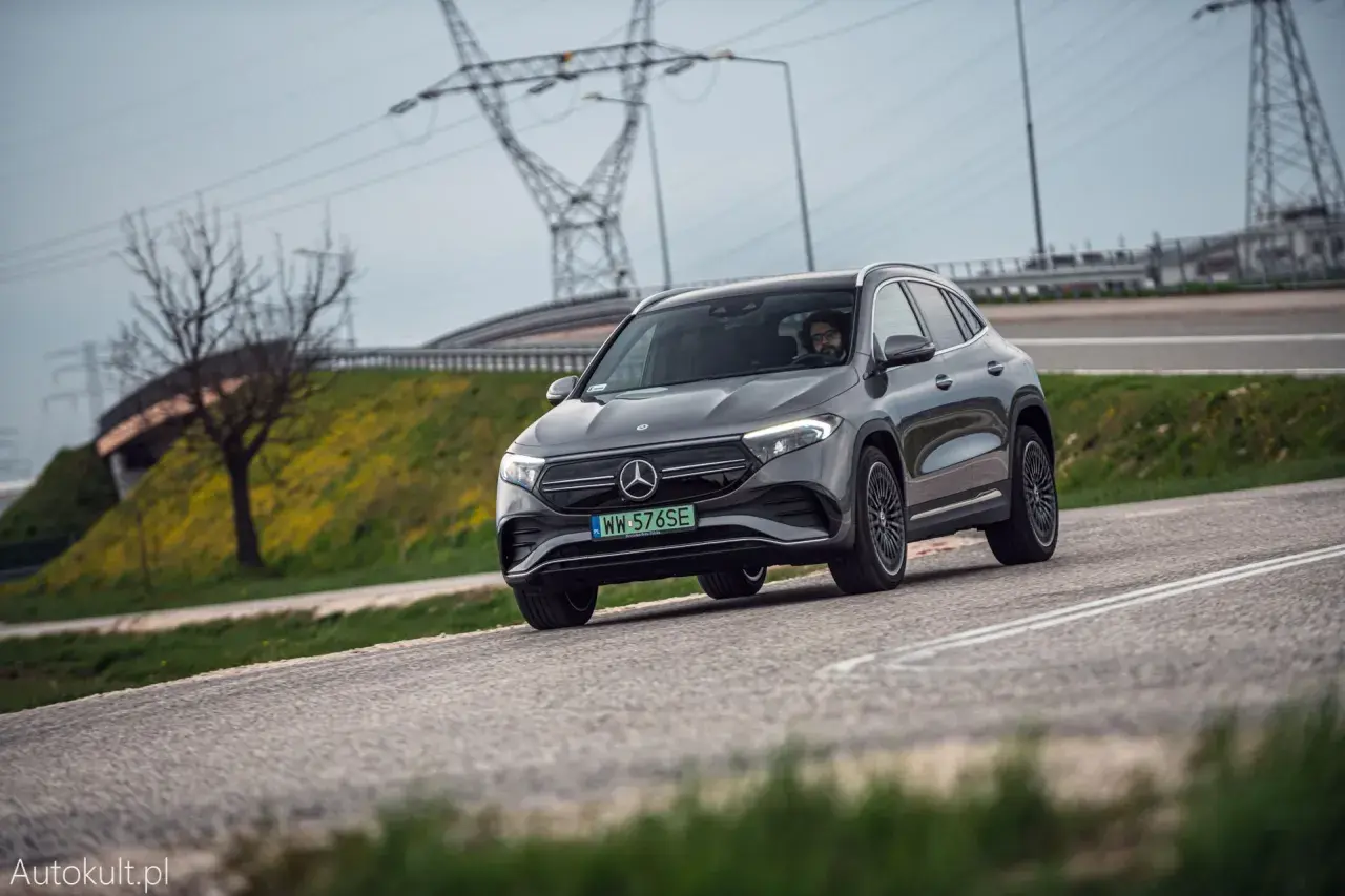 Szary Mercedes-Benz EQA jedzie po drodze, w tle linie energetyczne i drzewo.