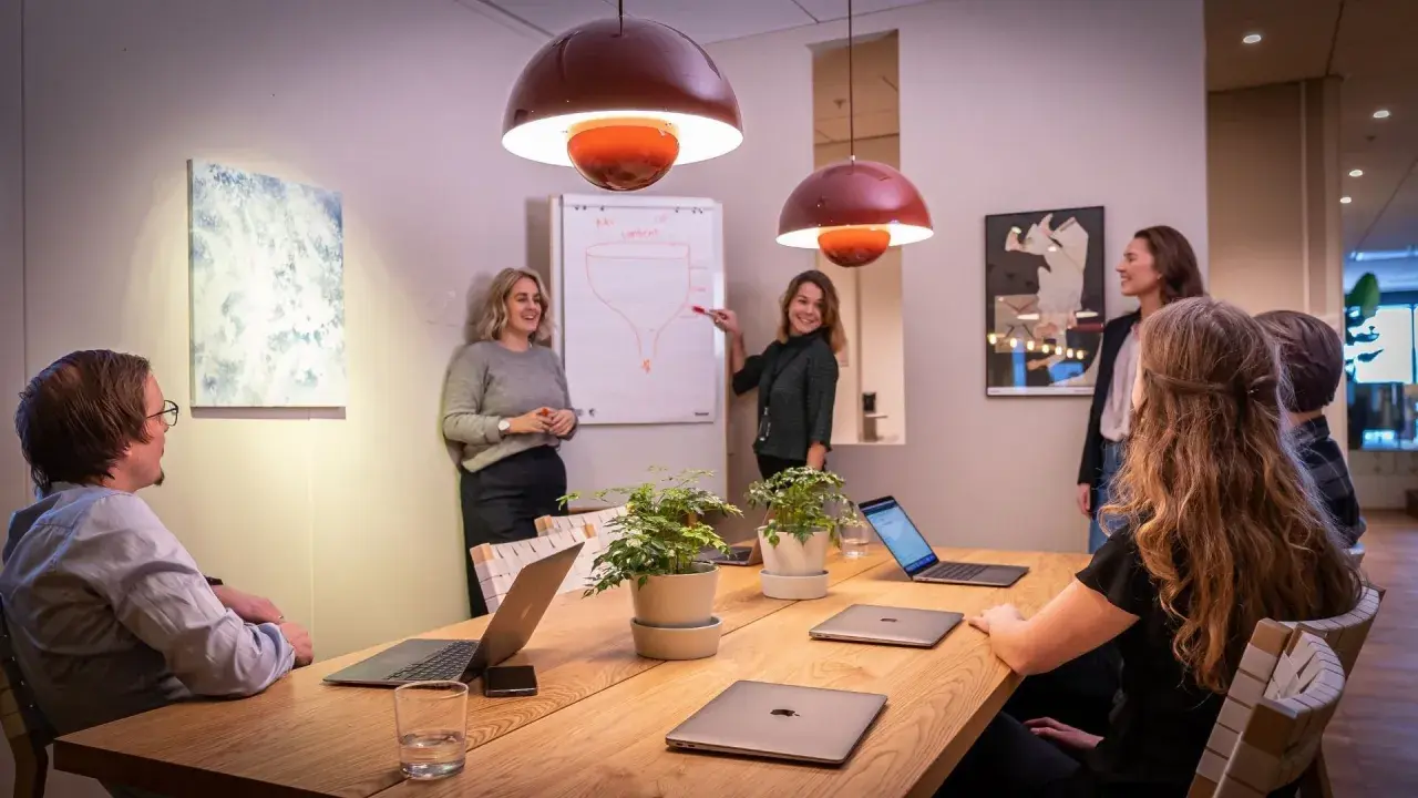 Ett team diskuterar strategier vid ett konferensbord med flera laptops. En kvinna pekar på en trattmodell på en whiteboard. Ups datorn är en del av deras effektiva arbetsflöde.