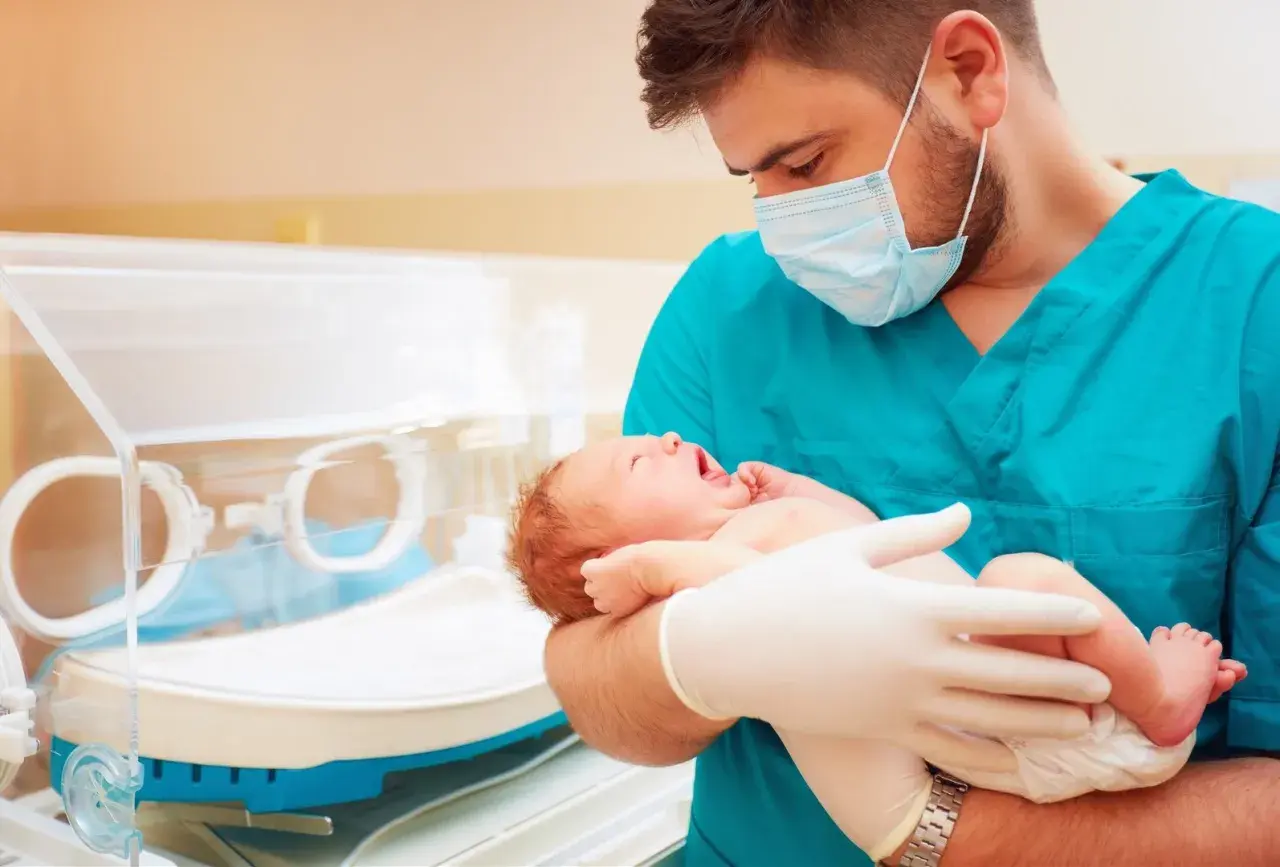 Lekarz w maseczce i rękawiczkach trzyma noworodka. To przykład troski w neonatologii, która zajmuje się opieką nad noworodkami.