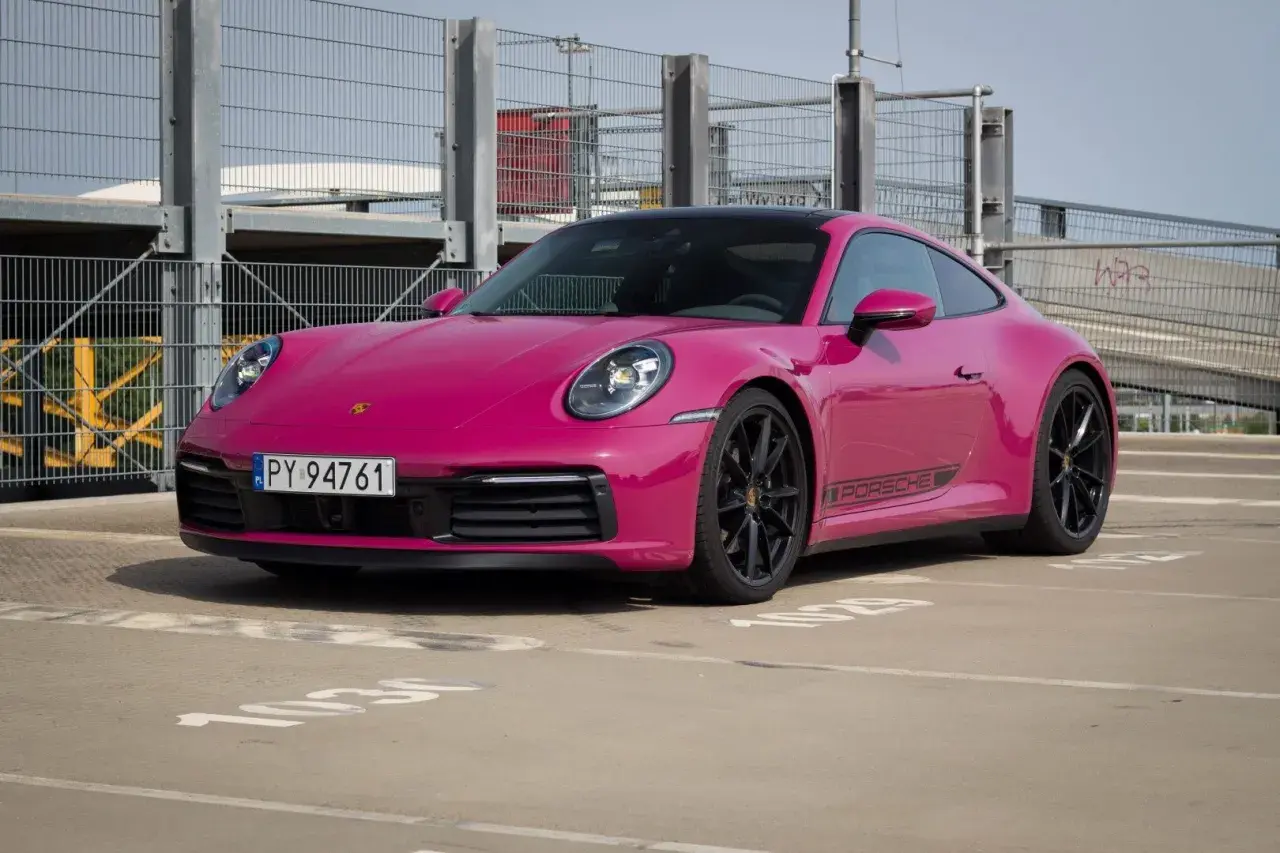 Różowy Porsche 911 Carrera 4S z czarnymi felgami i naklejką PORSCHE na progu, zaparkowany na zewnątrz.