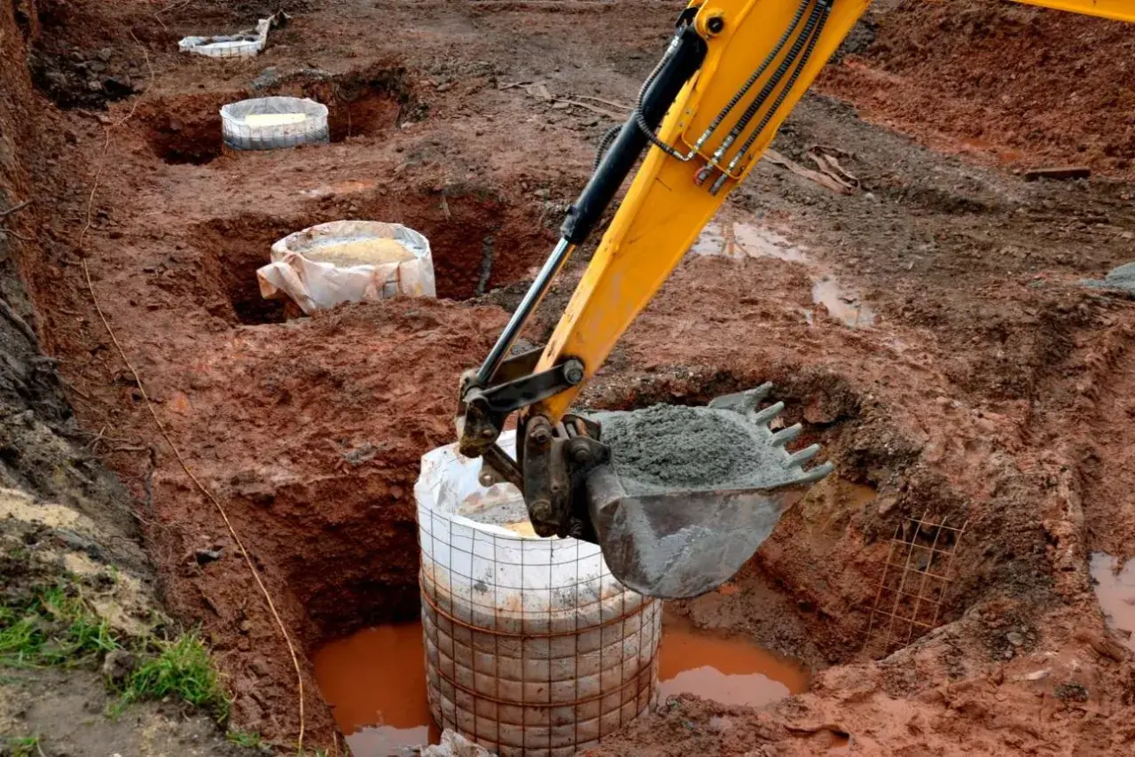 Koparka zasypuje betonem fundament palowy w wykopie. W tle widać kolejne przygotowane do zalania pale.