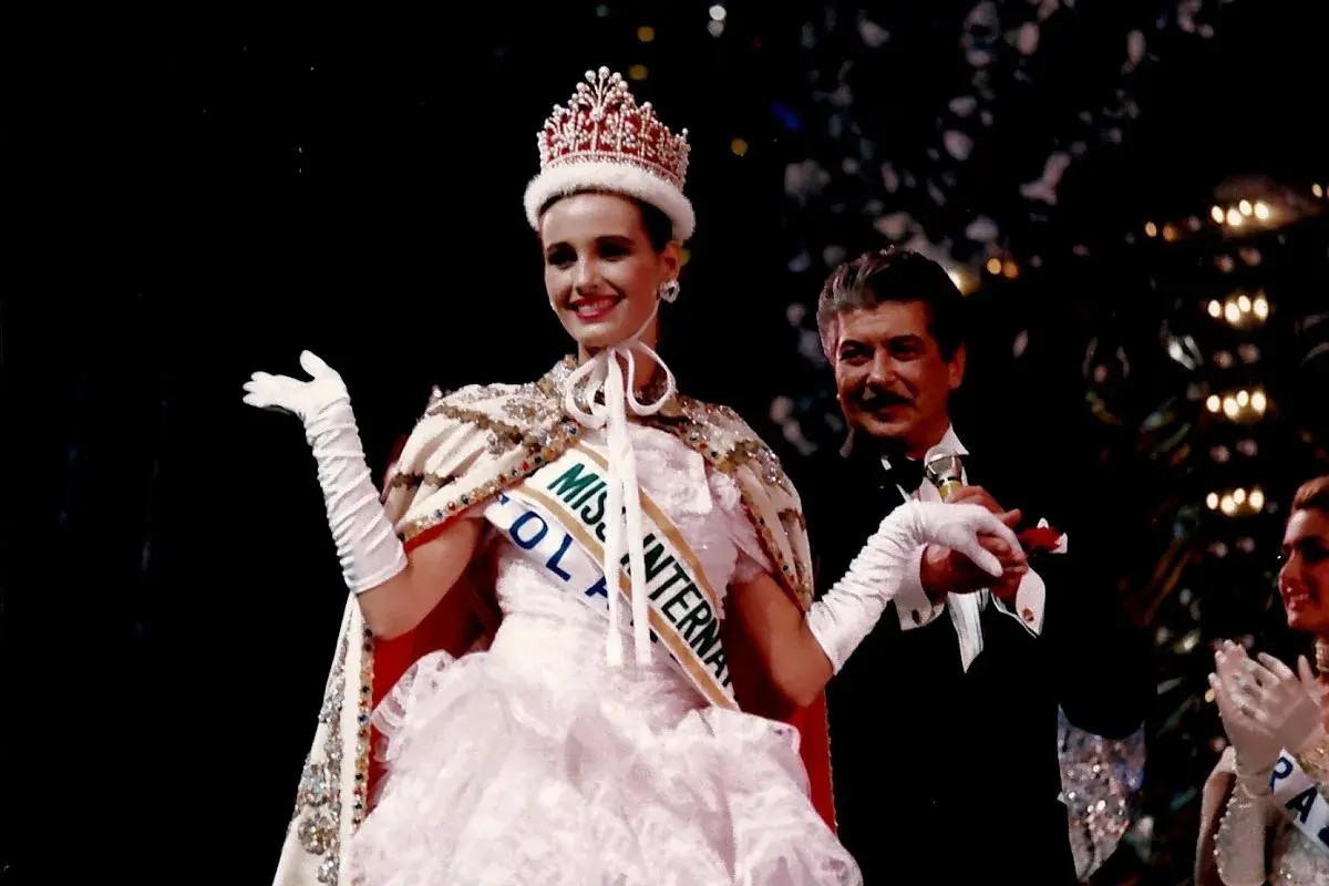 Uśmiechnięta Miss International w koronie i białej sukni, z szarfą "Miss Polonia". Obok niej mężczyzna w smokingu.