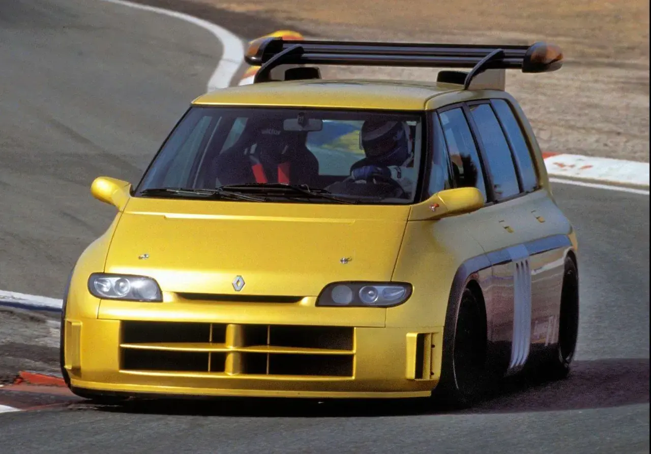 Żółty Renault Espace F1 z kierowcą w kasku na torze wyścigowym. Potężny spojler dachowy i agresywny wygląd.