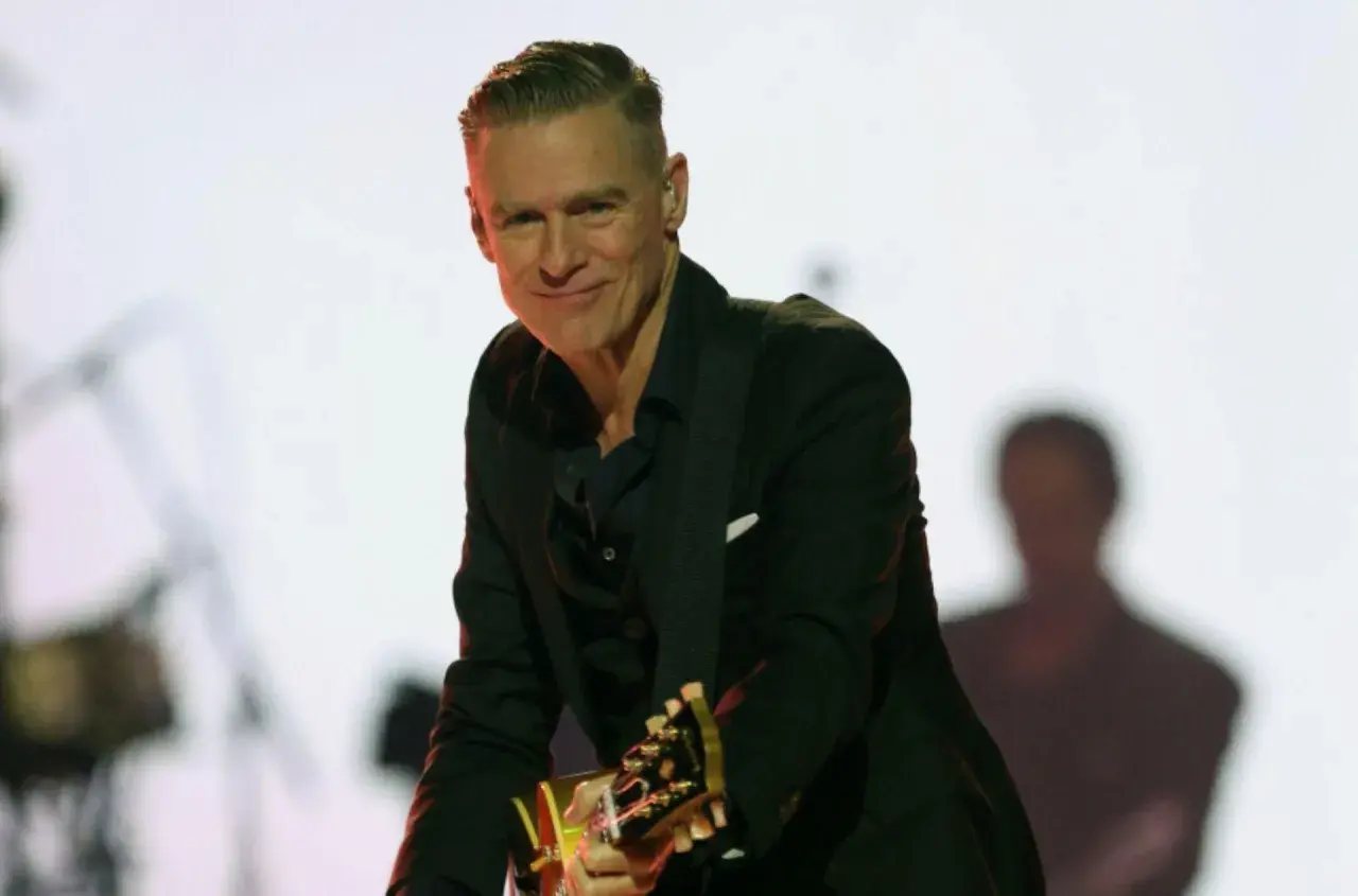 Bryan Adams w czarnym garniturze gra na gitarze. Sprawdź jego nadchodzące wydarzenia i koncerty.