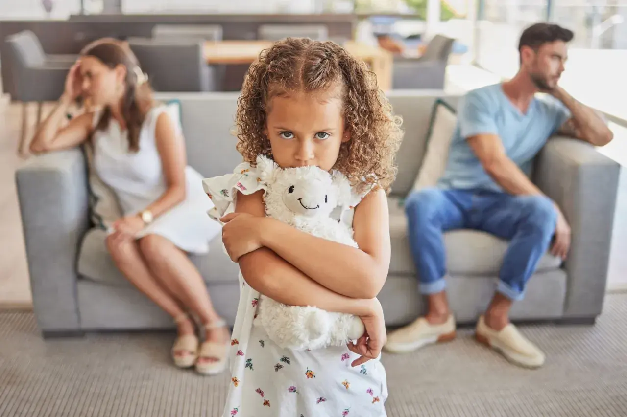 Ein Kind umklammert sein Stofftier, während die Eltern im Hintergrund getrennt sitzen. Ein Bild, das die emotionale Belastung eines Trennungsjahres mit Kindern widerspiegelt.