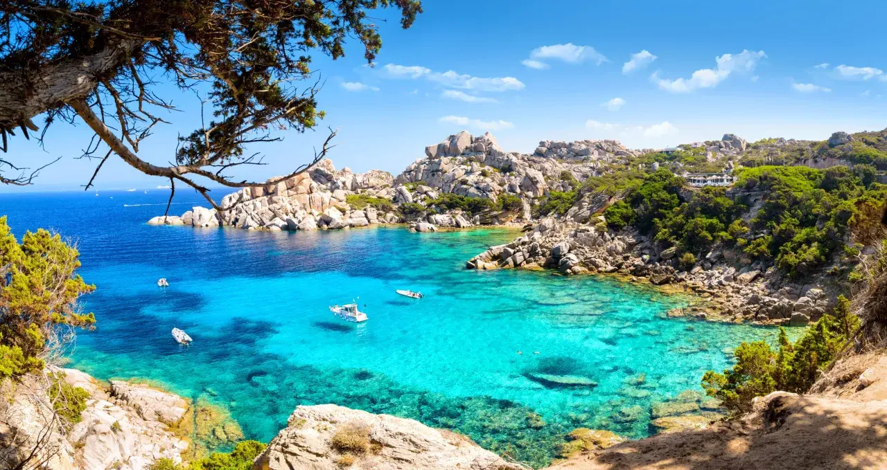 Turkusowa woda i skaliste wybrzeże Costa Smeralda na Sardynii. Kilka łodzi kołysze się na falach.