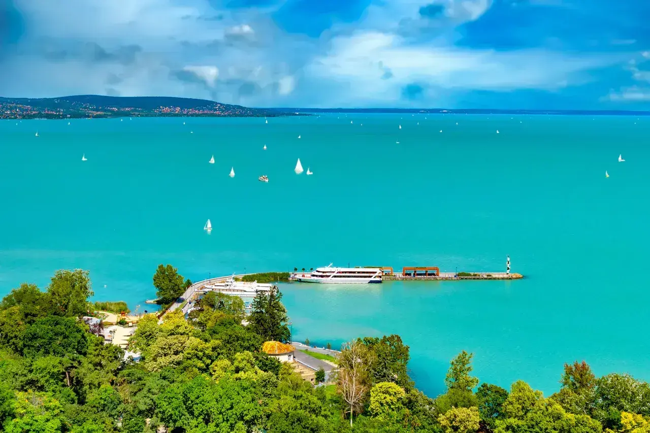 Turkusowe wody jeziora Balaton, pełne żaglówek i statków, z zielonymi wzgórzami w tle.