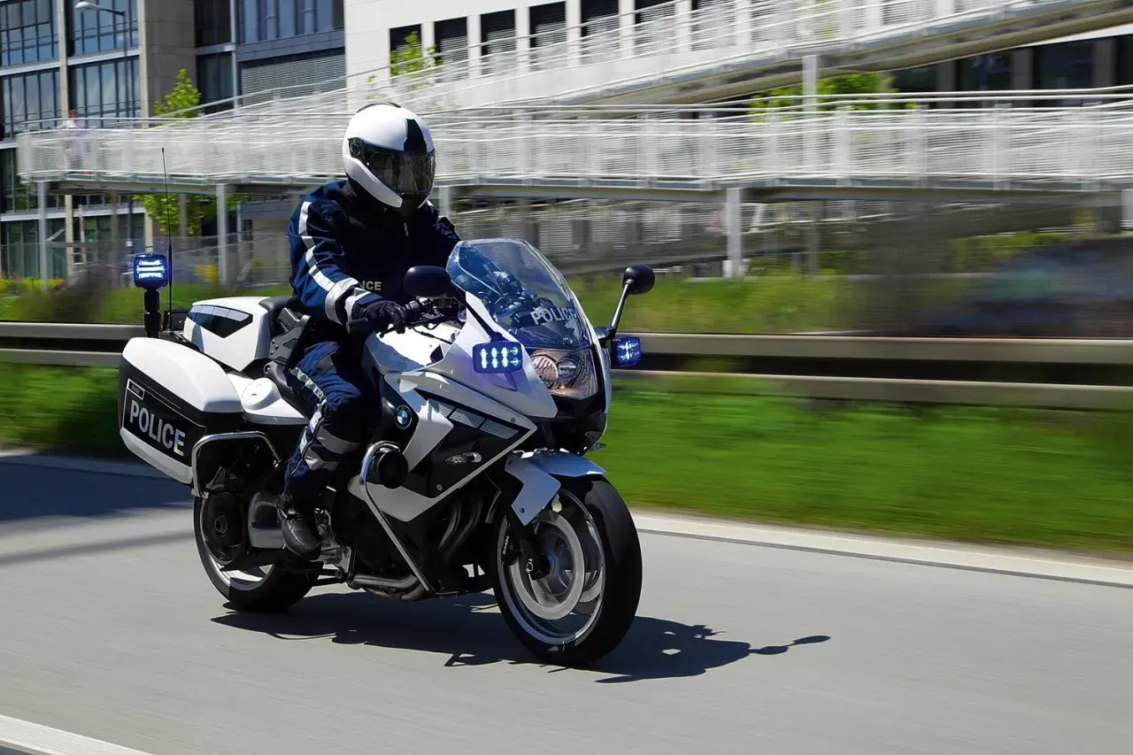 Policyjny motocykl BMW F800GT z włączonymi sygnałami świetlnymi pędzi po drodze.