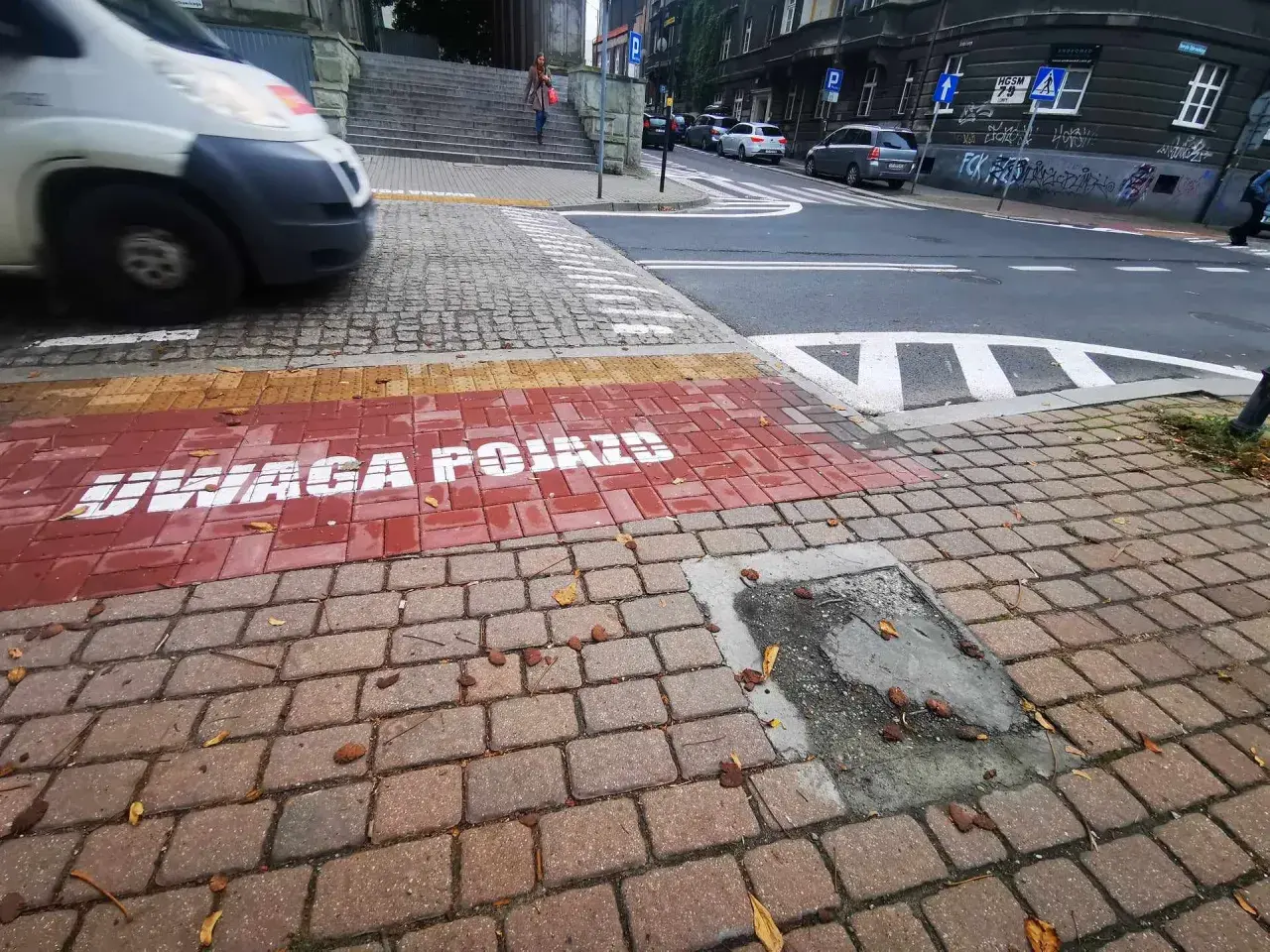 Czerwony pas na chodniku z napisem "UWAGA POJAZDY" informuje o przejściu sugerowanym. W tle widać samochody i schody.