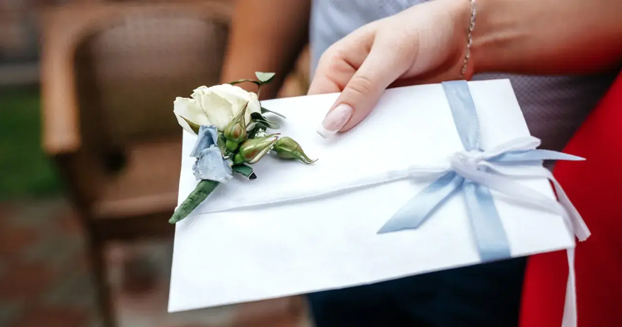 Dłoń trzyma białą kopertę z błękitną wstążką i ozdobnym kwiatkiem. Rozważasz skutki wręczenia pustej koperty lub zbyt małej kwoty? Opinie i etyka są tu kluczowe.