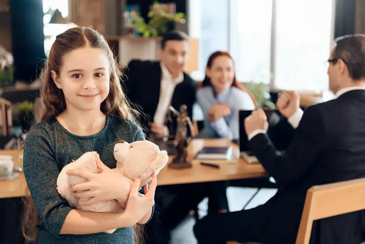 Uśmiechnięta dziewczynka z misiem w ramionach, podczas gdy rodzice i prawnik omawiają zmianę nazwiska dziecka.