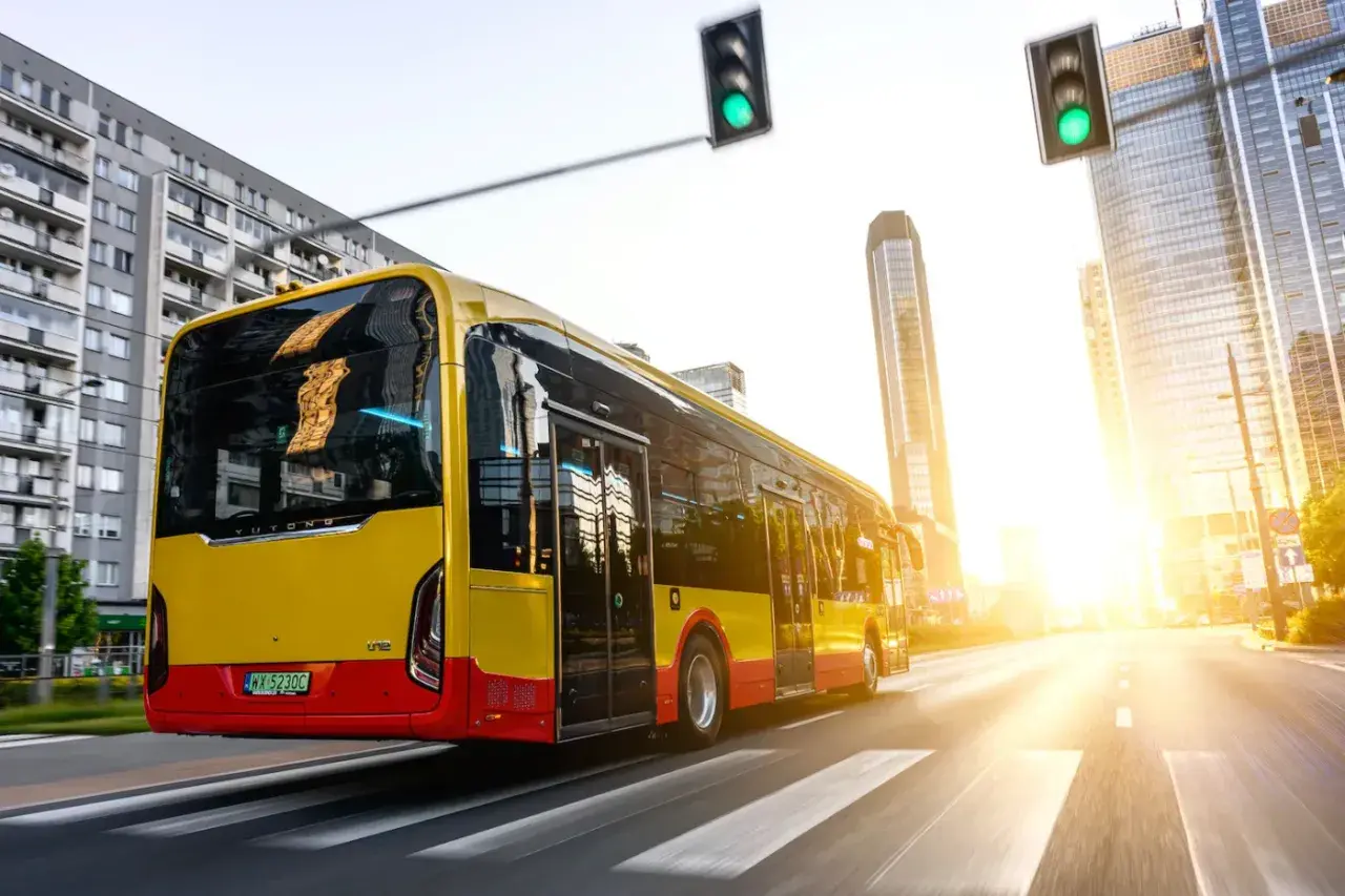 Żółto-czerwony autobus elektryczny Yutong jedzie przez miasto w blasku zachodzącego słońca.