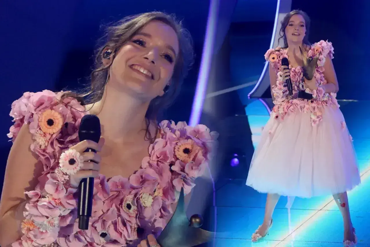 Zuzanna Irena Jurczak w kwiecistej sukni na scenie, trzyma mikrofon i uśmiecha się do publiczności.