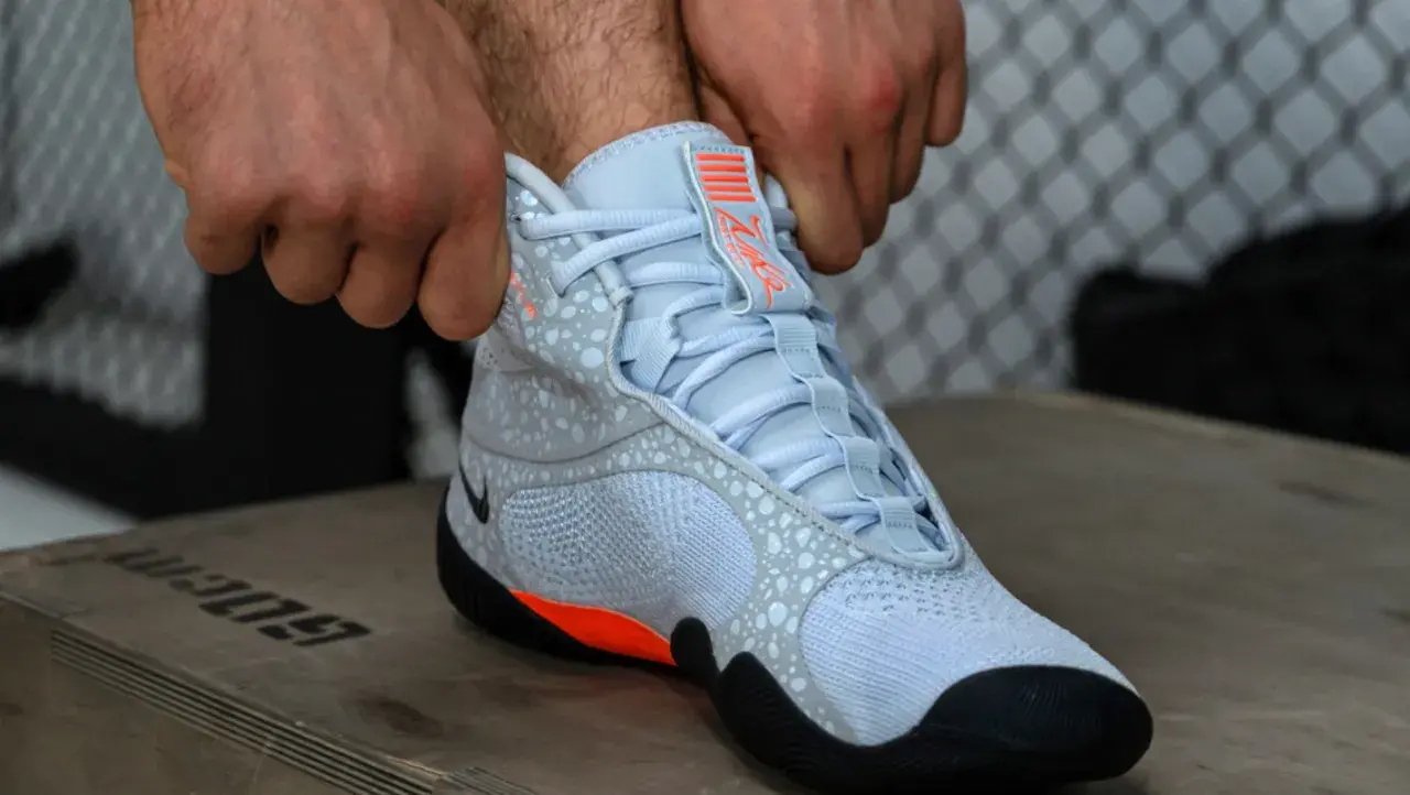 Ręce zapinają jasnoniebieskie buty sportowe z pomarańczowymi akcentami, przypominające styl Batistini.
