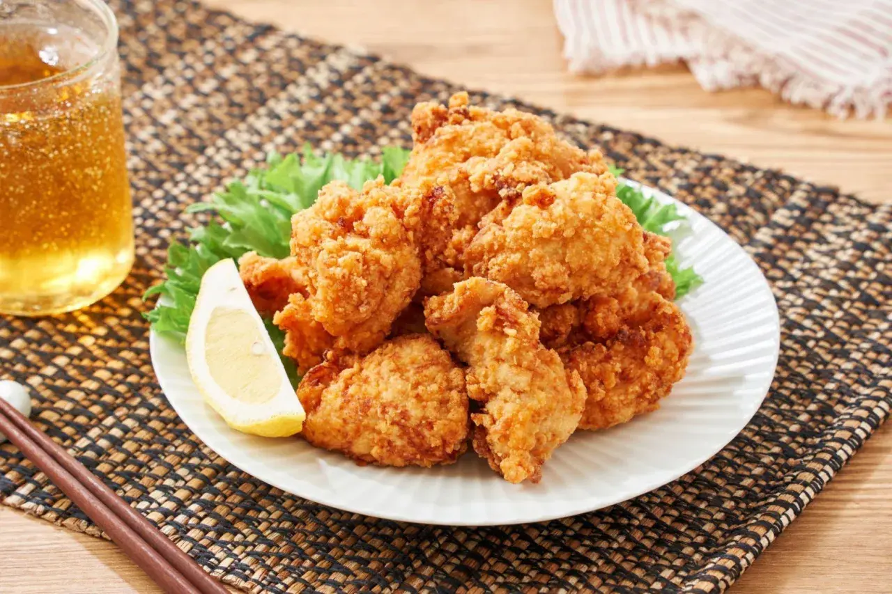 Chrupiące karaage chicken na talerzu, ozdobione plasterkiem cytryny i liśćmi sałaty. Obok szklanka złocistego napoju i pałeczki.