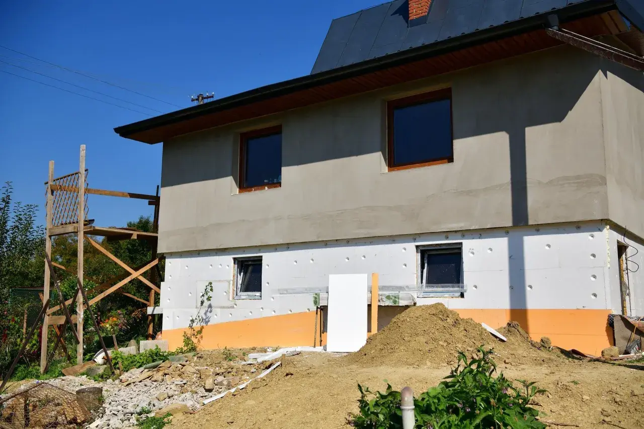 Budynek w trakcie ocieplania styropianem. Widoczne okna, rusztowanie i ziemia. Trzeba uważać na **mostek termiczny** przy oknach.