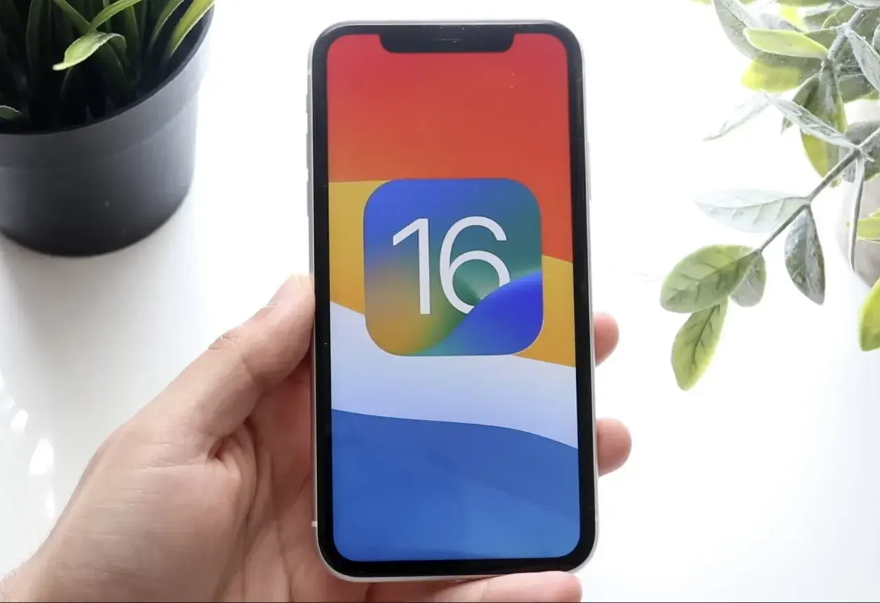 Dłoń trzyma iPhone'a z ikoną iOS 16 na kolorowej tapetcie.
