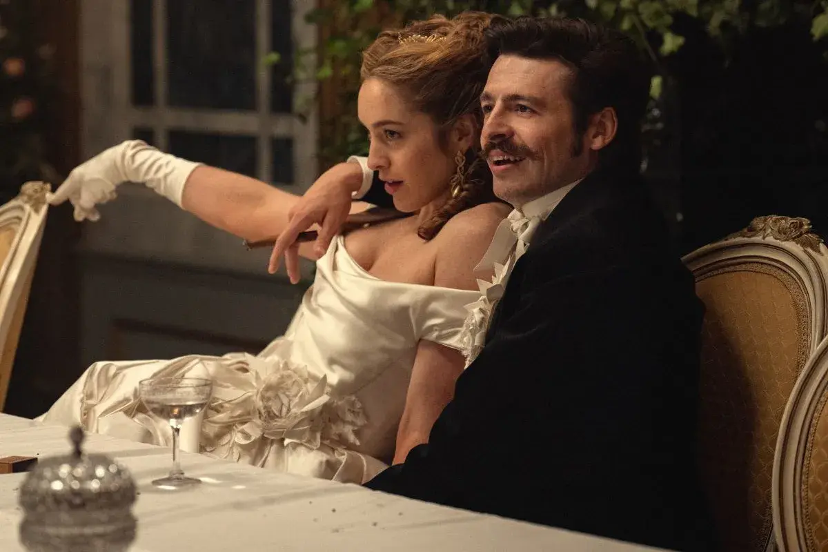 Elegancka para przy stole, zapowiedź nowych produkcji Netflix. Kobieta w białej sukni z rękawiczkami, mężczyzna w czarnym fraku.