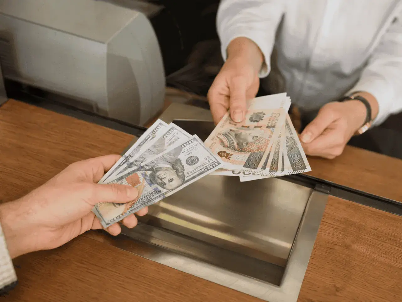 Dwie ręce wymieniają banknoty. Jedna trzyma banknoty 100 dolarów, druga polskie 200 złotych.