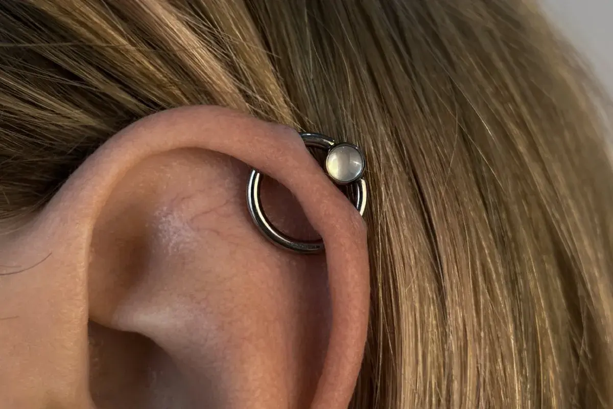 Oryginalny kolczyk orbital piercing z białym kamieniem na uchu, otoczony blond włosami.